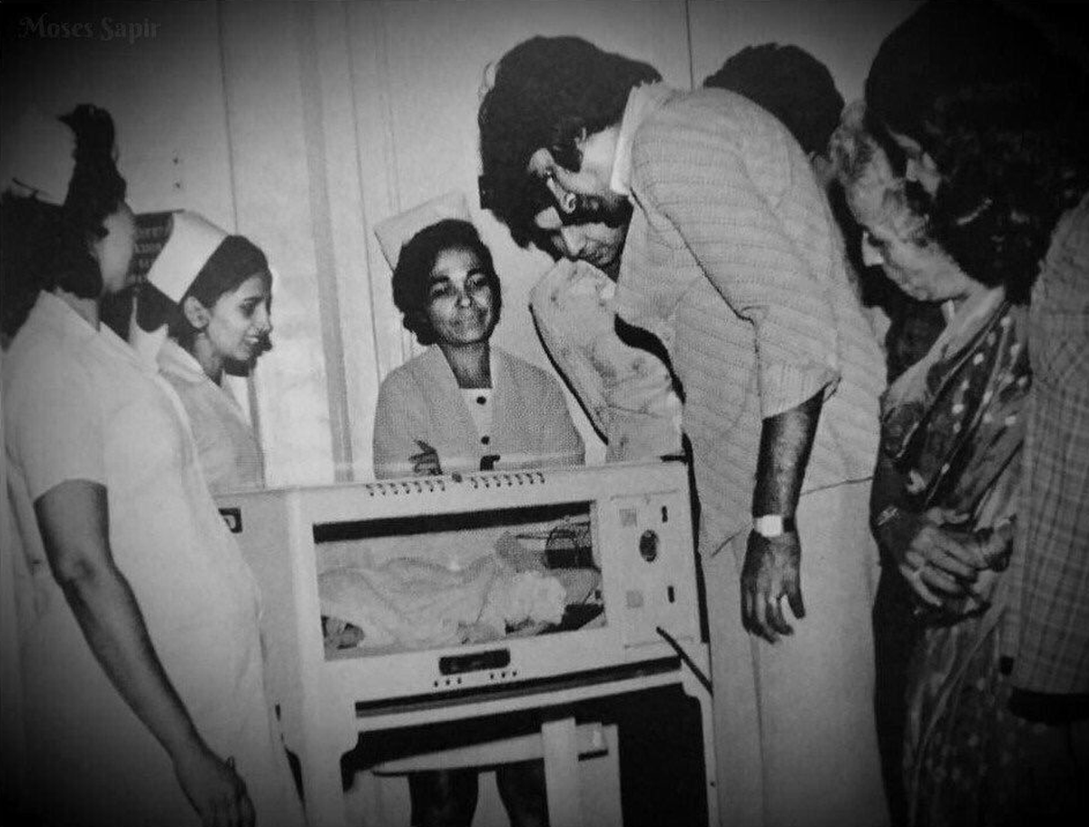 Amitabh in the hospital is surrounded by nurses near an infant incubator, who is affectionately staring at the newborn Abhishek.