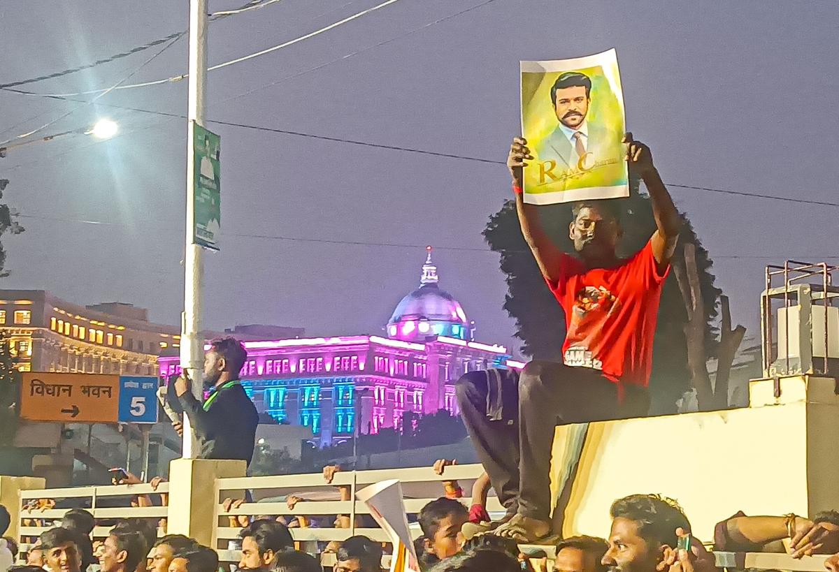 A fan of Ram Charan climbs on top of a parapet wall at Pratibha Theatre in Lucknow, with an illuminated Vidhan Sabha in the background