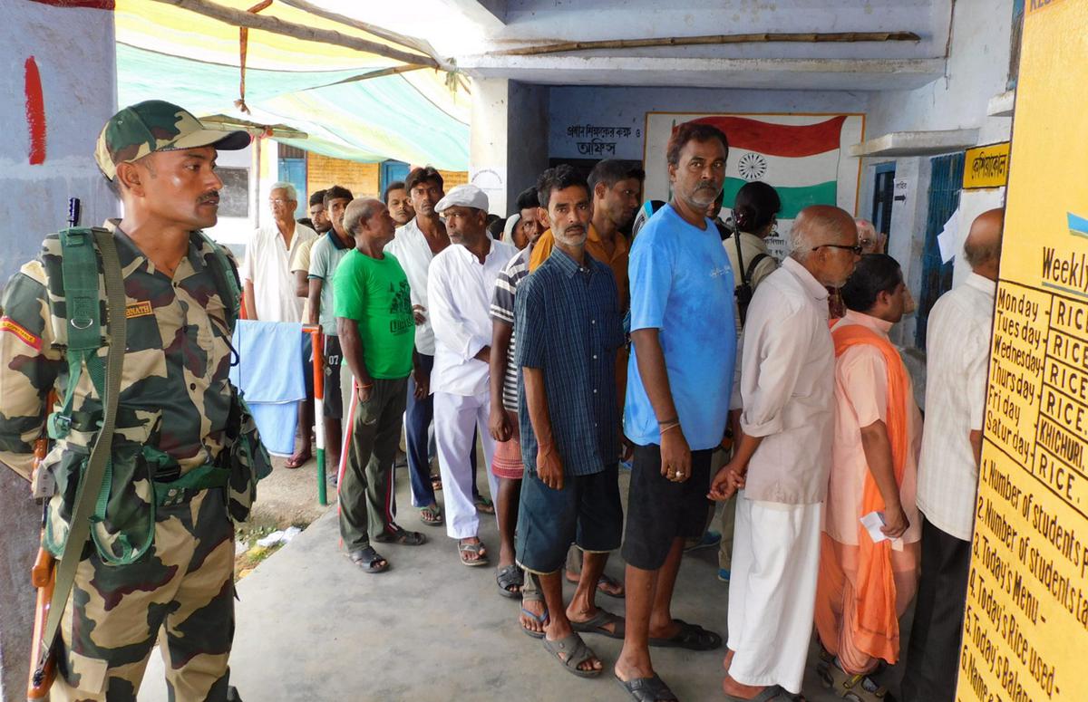 A security person stands guard as people stand in a queue to cast their votes for Panchayat elections at a polling station, in Bankura, Saturday, July 8, 2023. 