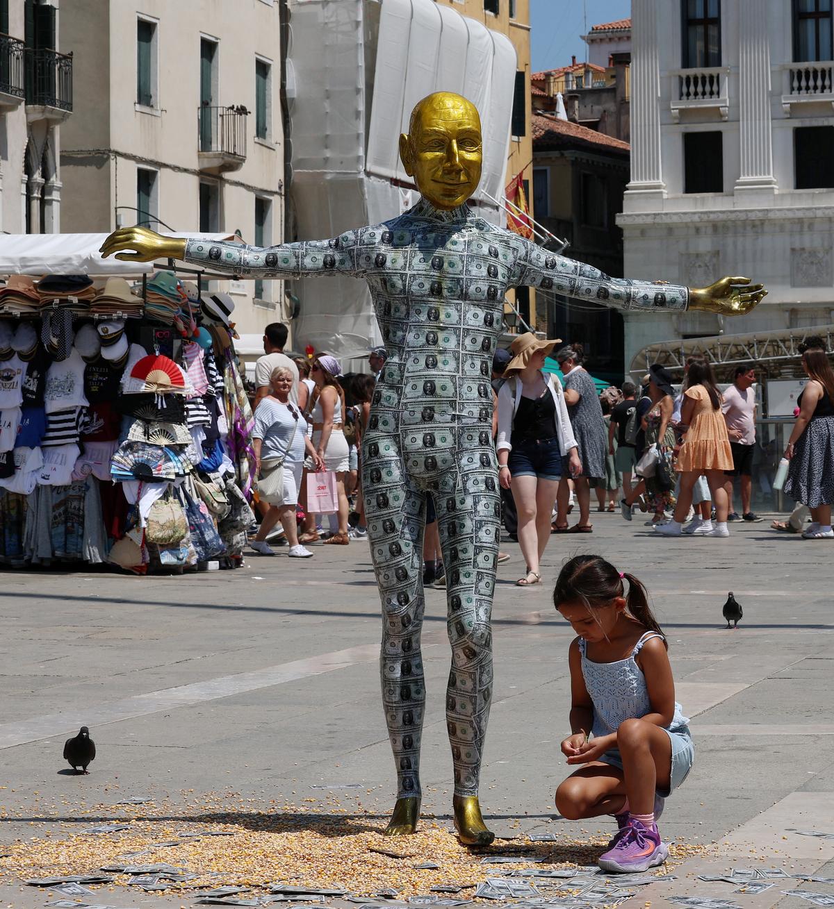 A child kneels next to a mannequin depicting Amazon Founder Jeff Bezos covered in fake dollar bills installed at St. Mark’s Square as part of a parody stunt by art collective Konn Artiss, ahead of the wedding in Venice