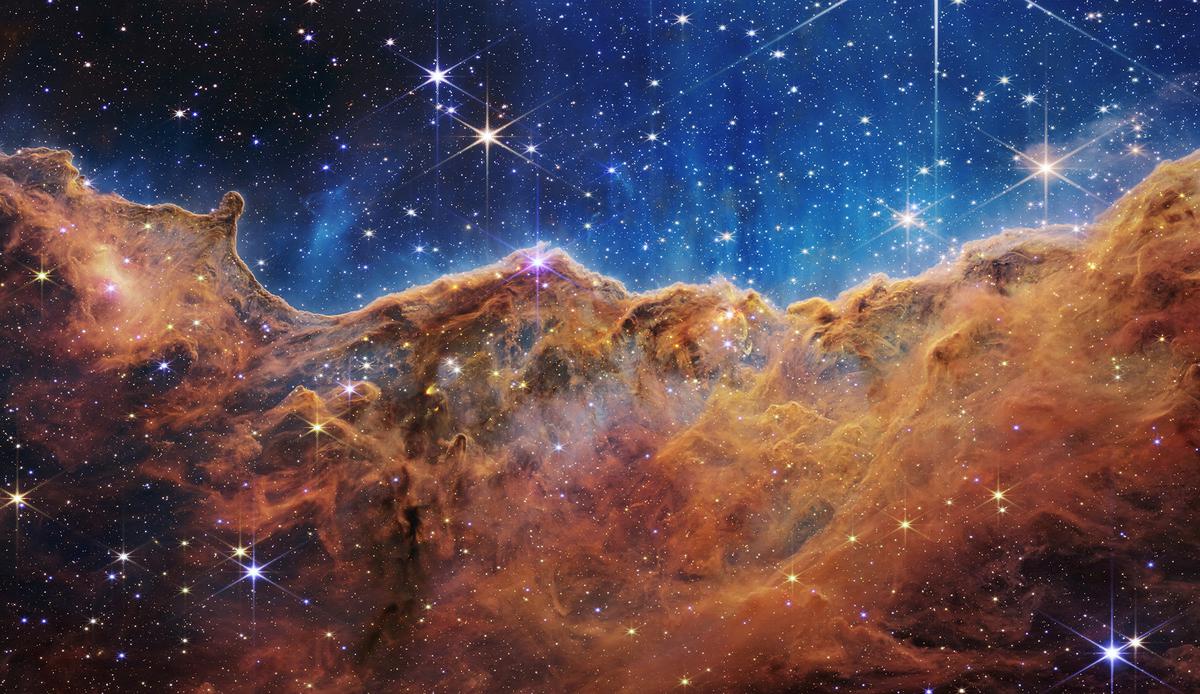 The “Cosmic Cliffs” of the Carina Nebula is seen in an image divided horizontally by an undulating line between a cloudscape forming a nebula along the bottom portion and a comparatively clear upper portion, with data from NASA’s James Webb Space Telescope.