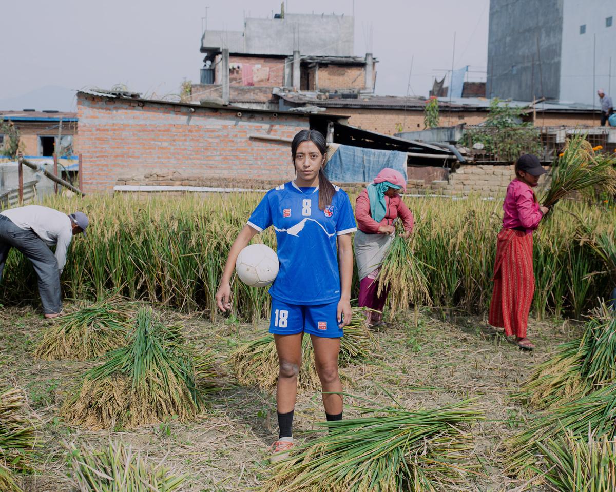 Photograph by Amit Machamsi; A football player poses for the camera amidst the bustling scene of farmers harvesting paddy in Sipadol, Bhaktapur. 
