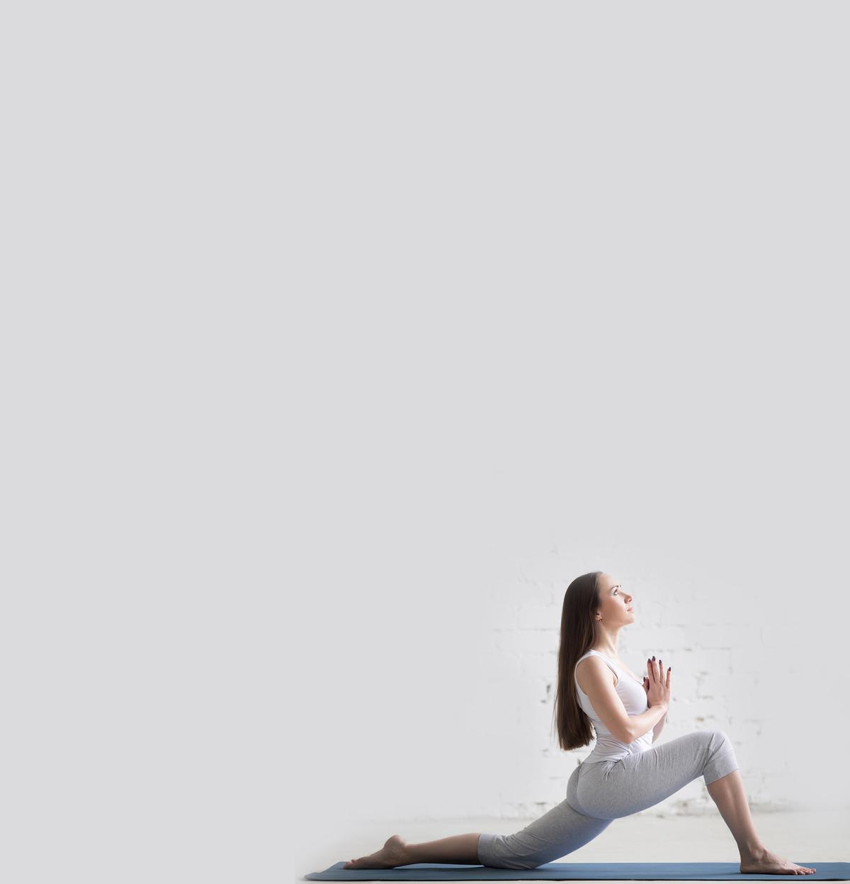 Attractive happy young woman working out indoors. Beautiful model doing exercises on blue mat in room with white walls. Standing in low lunge exercise, anjaneyasana. Full length. Side view