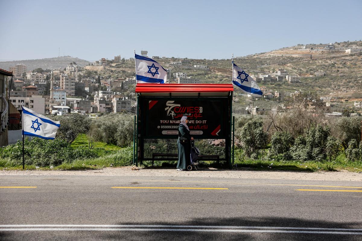 A Palestinian woman waits at a bus stop next to Israeli flags, near Luban e-Sharkiya, between Nablus and Ramallah, in the Israeli-occupied West Bank, on February 16, 2026. 