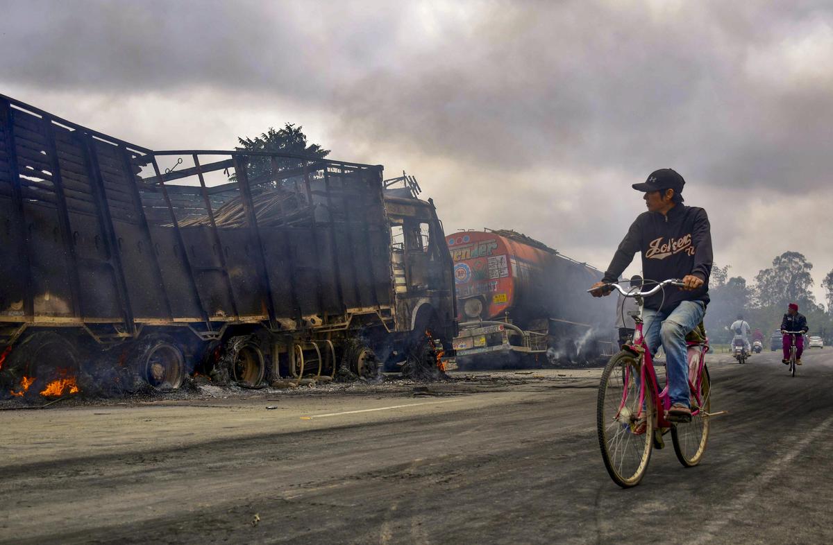 7 अप्रैल, 2026 को मणिपुर के बिष्णुपुर जिले में एक बम हमले में दो बच्चों की मौत और उनकी मां के घायल होने के विरोध में लोगों द्वारा जलाए गए ईंधन टैंकरों के जले हुए अवशेषों के पास से गुजरते लोग।