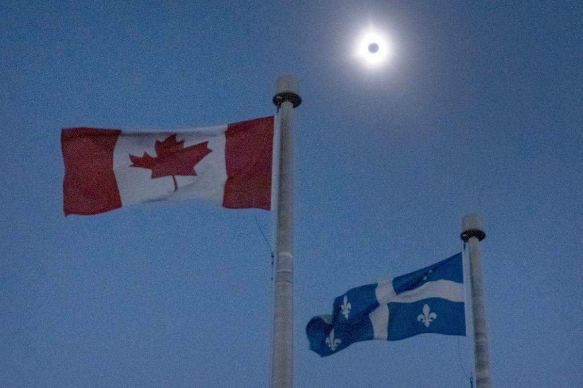 The moon covers the sun during a total solar eclipse, as Canadian and Quebec flags fly, as seen from Bishop’s University in Sherbrooke, Quebec, Monday, April 8, 2024.