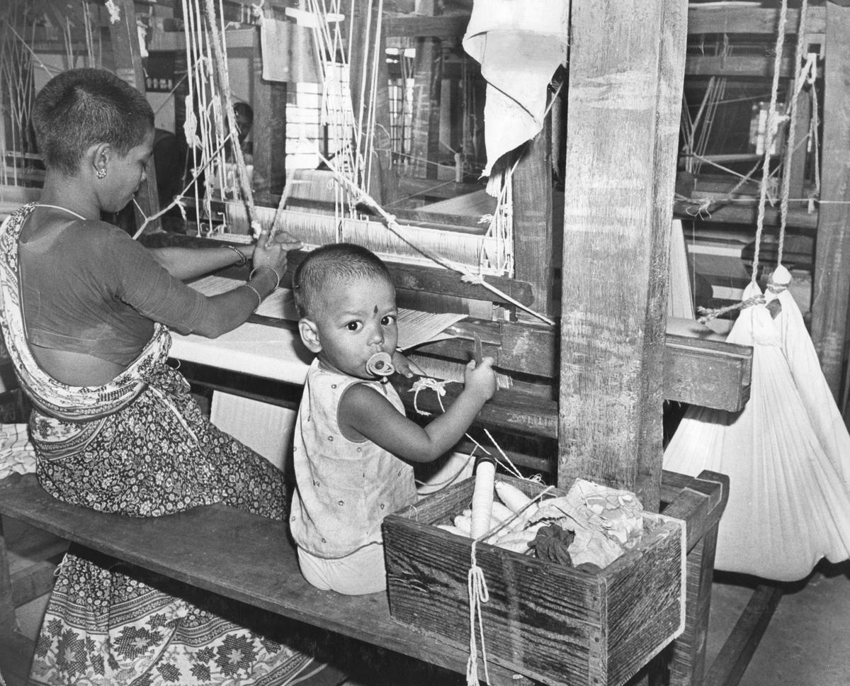 Seorang wanita, dengan anaknya di dekatnya, terlihat sedang bekerja di bagian tenun masyarakat koperasi Pondicherry State Weavers pada tahun 1982 Seorang wanita, dengan anaknya di dekatnya, terlihat sedang bekerja di bagian tenun masyarakat koperasi Pondicherry State Weavers pada tahun 1982