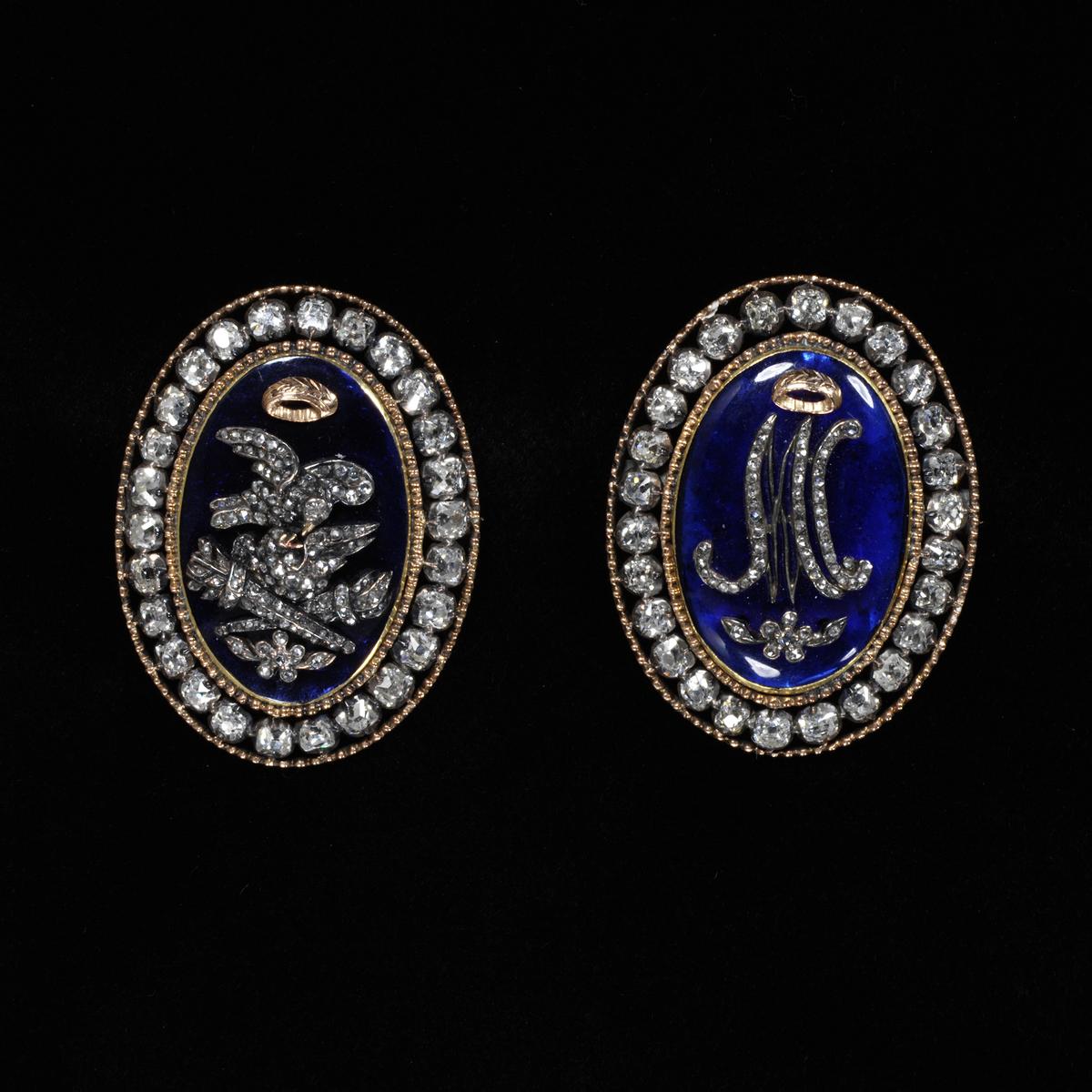 A pair of braided bracelet clasps; gold with brilliant-cut diamonds with central plaques of blue paste, one with the initials of Marie Antoinette, and the other with turtle doves and hymeneal torches. (Paris, 1770)