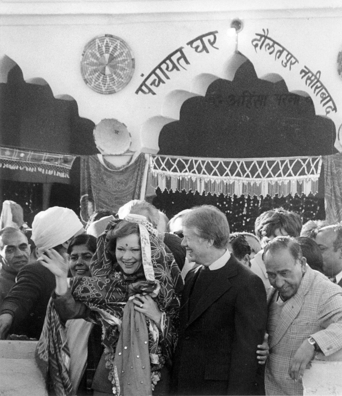 Rosalynn Carter wears a shawl presented to her by the villagers in Daulatpur Nasirabad, India, as President Carter smiles approvingly. The spot of red dye on the forehead, a tilak, is a Hindu mark of respect and friendship. The name of the village was permanently changed to Carterpuri in honour of their visit.