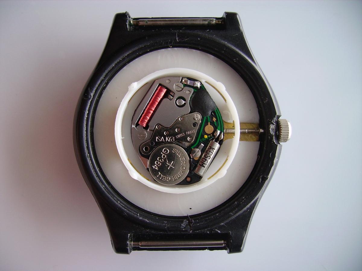 The clockwork of a basic quartz wristwatch. Bottom right: quartz crystal oscillator, left: button cell watch battery, top right: oscillator counter, digital frequency divider and driver for the stepping motor (under black epoxy), top left: the coil of the stepper motor that powers the watch hands.