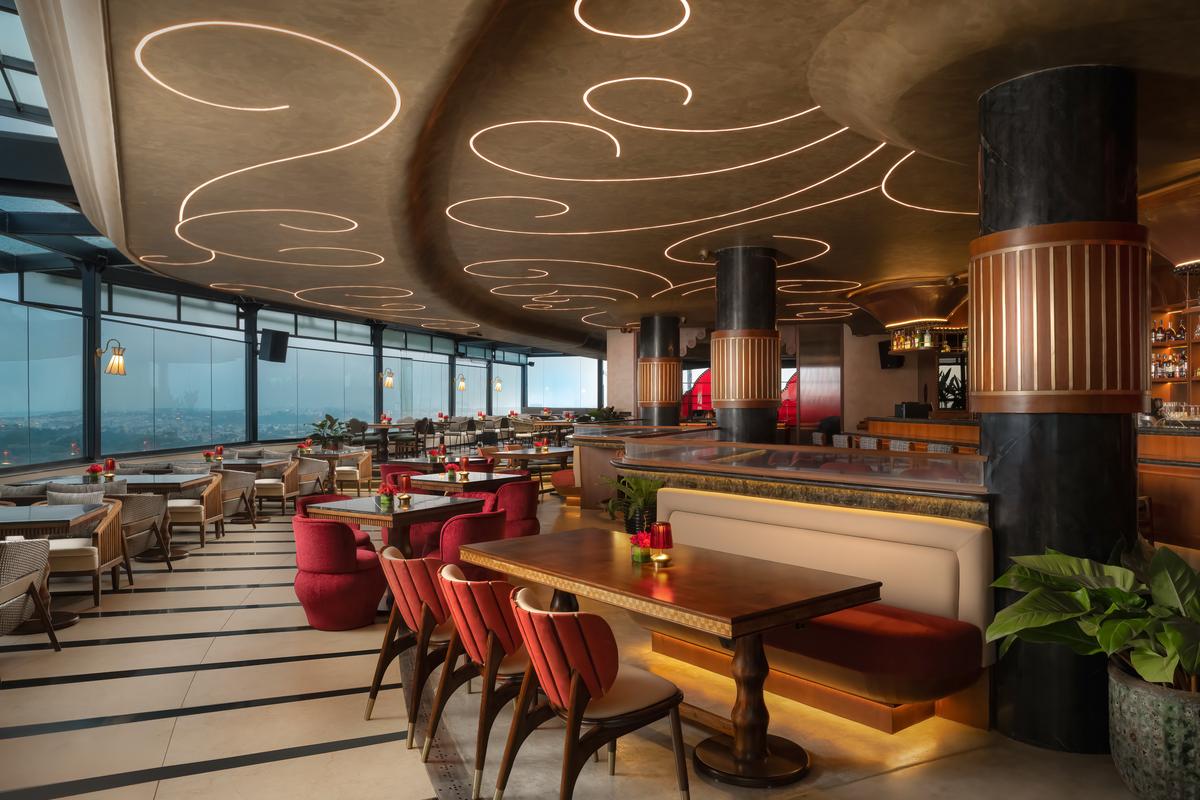 Sculptural ceiling lines ripple across the dining space, framing warm seating against the Bengaluru skyline. Sculptural ceiling lines ripple across the dining space, framing warm seating against the Bengaluru skyline.